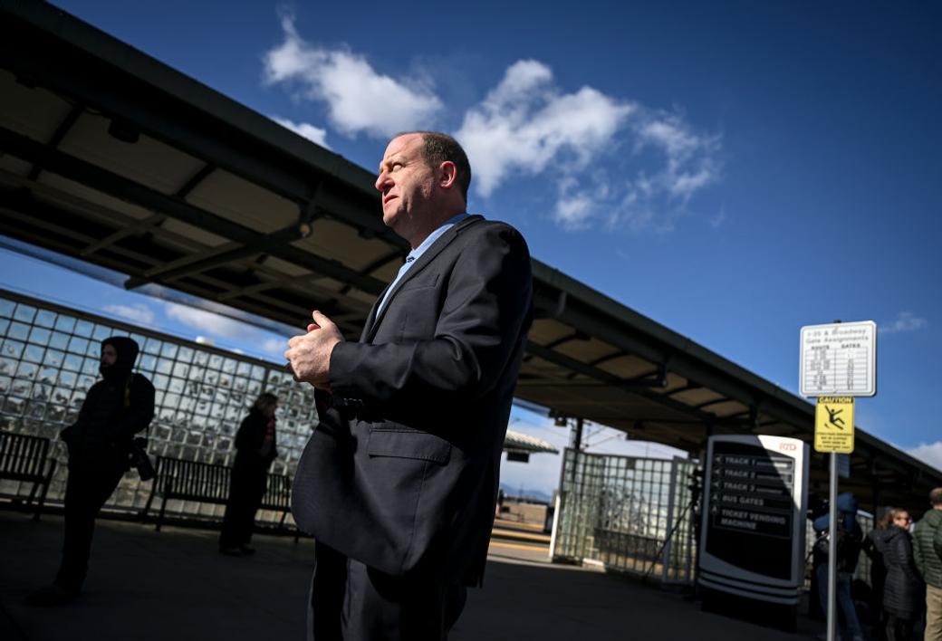 Polis stands outside an RTD light rail stop in 2024