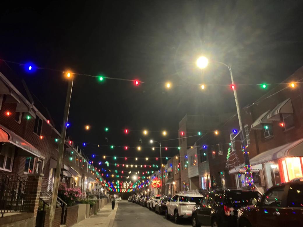 A residential street with row homes on either side is lit up by multicolored string lights hanging above the street. A street lamp is brightly lit, with multicolored string lights wrapped around its base. Several other homes are illuminated by Christmas-themed light decorations. 