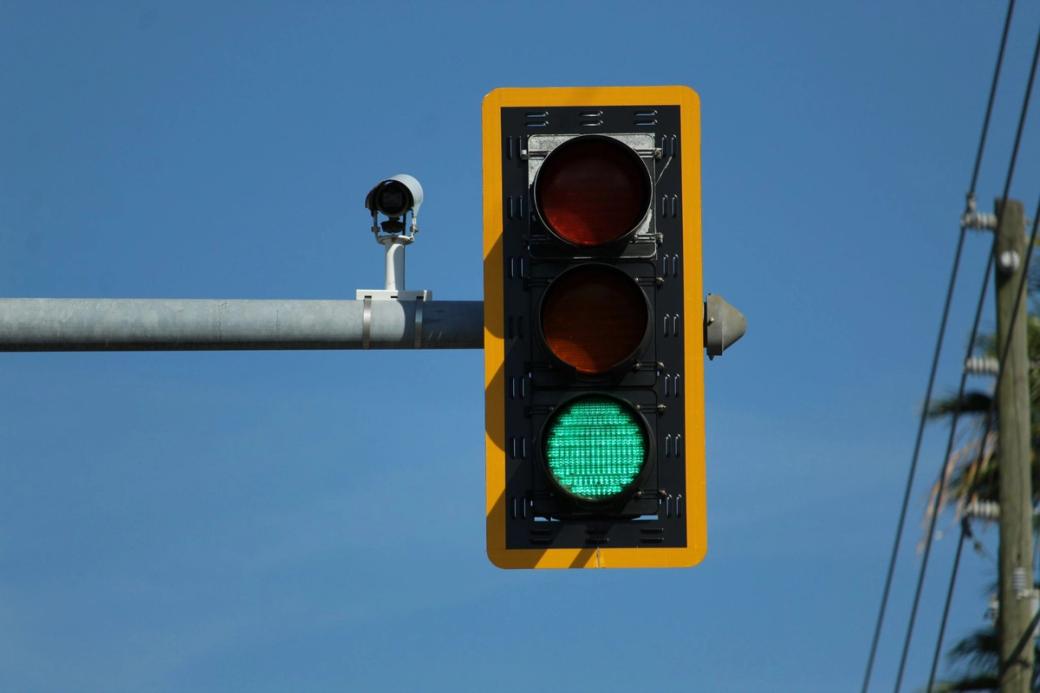 A traffic light showing green, hanging from a horizontal pole with a camera on it.