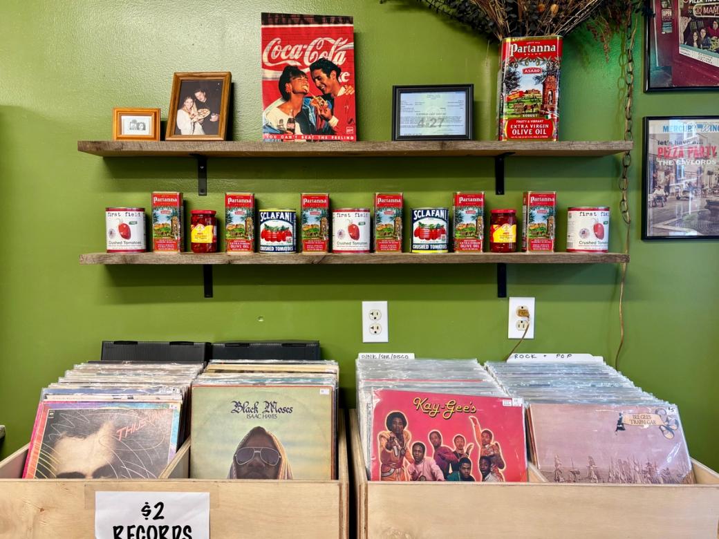 The interior of a pizza shop with an olive green wall displaying two wooden shelves lined with cans of Partanna olive oil, Sclafani crushed tomatoes, and other Italian pantry goods, along with framed photos and a vintage Coca-Cola advertisement. Below, wooden crates hold bins of vinyl records for $2, with visible albums including Isaac Hayes' 'Black Moses' and Kay-Gee's, sorted by genres including Funk/Soul/Disco and Rock & Pop.