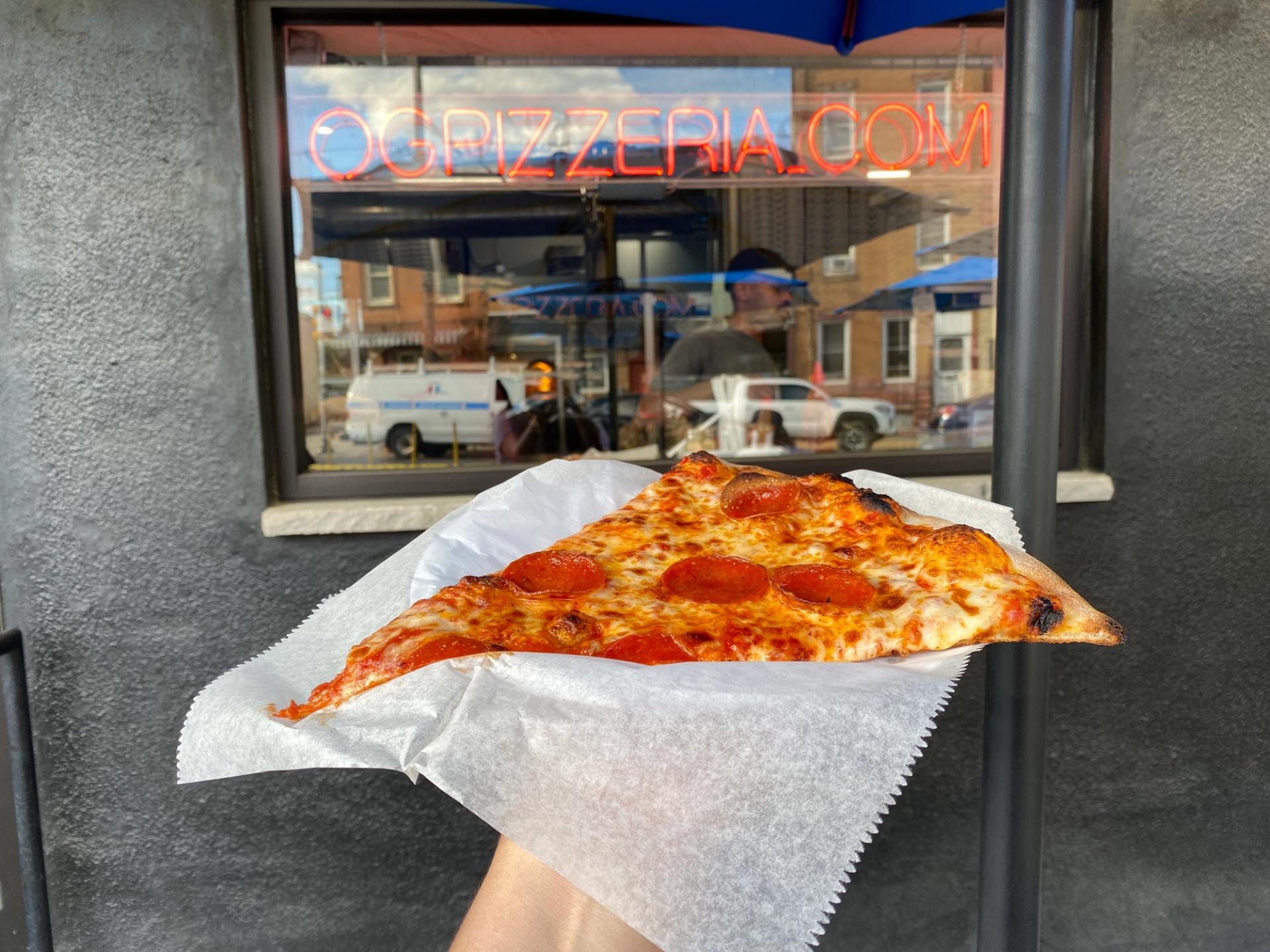 A slice of pepperoni pizza in front of a sign that says "OGPIZZERIA.COM"
