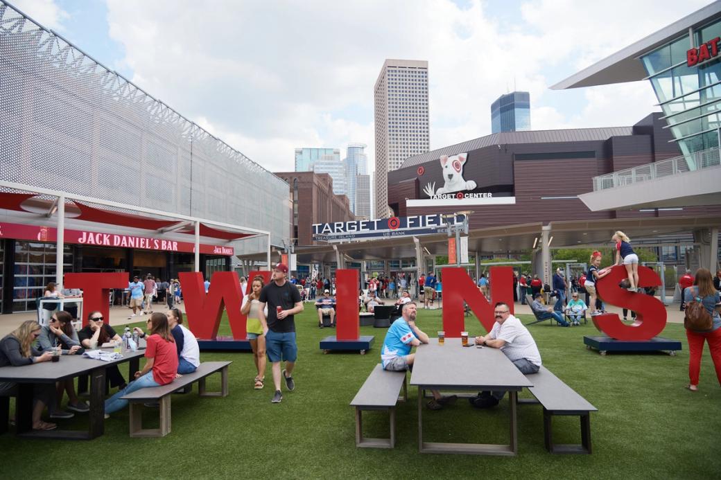 Lots of people on Target Field, with red letters spelling out "Twins." Two picnic tables are in the foreground, with tall buildings outside the stadium in the background.