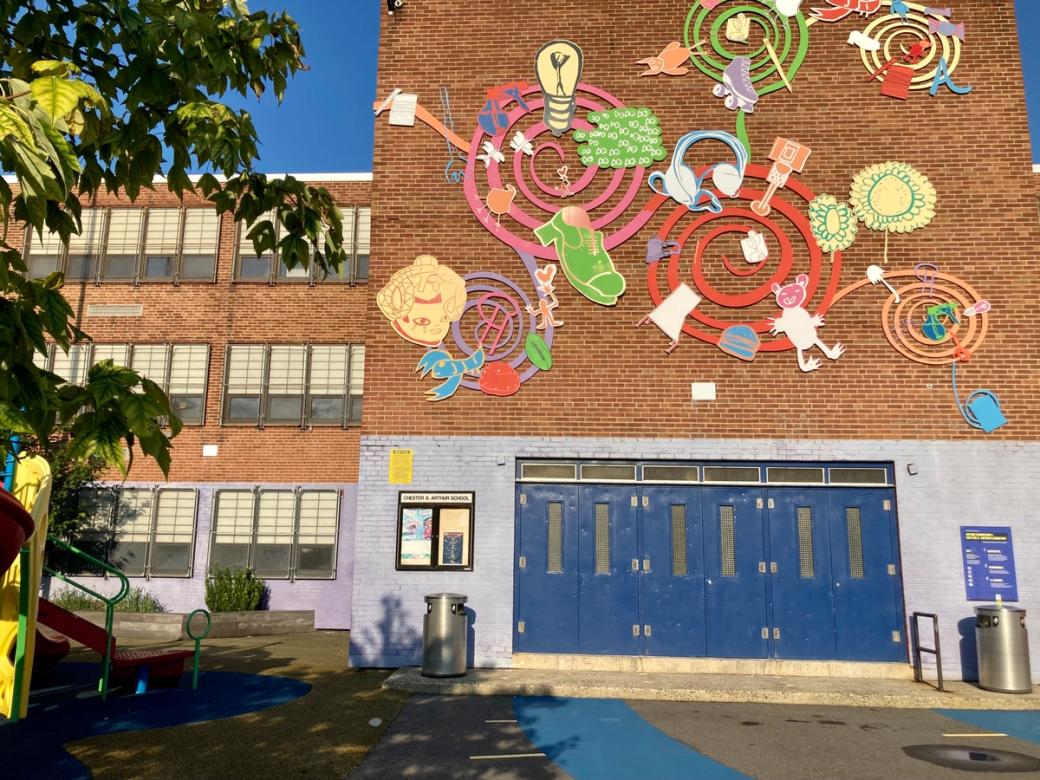 The exterior of Chester A. Arthur School, a red brick elementary school building, decorated with a large colorful mural of swirling spirals and whimsical cut-out shapes including a lightbulb, sneaker, headphones, figures and flowers. A playground and blue entry doors are visible at ground level.