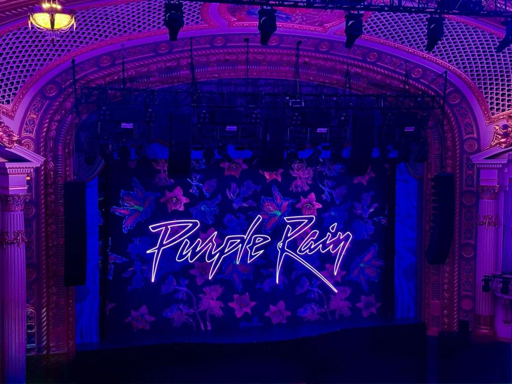 Empty stage lit in shades of purple with stylized font on large banner that reads, "Purple Rain"