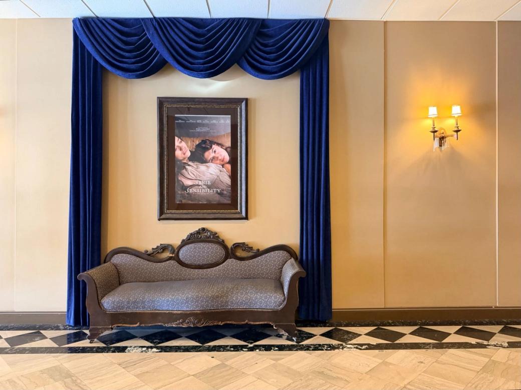  An ornate vintage sofa sits beneath deep blue velvet drapes inside the Manor Theater, with a framed “Sense and Sensibility” poster above it and a glowing wall sconce nearby.