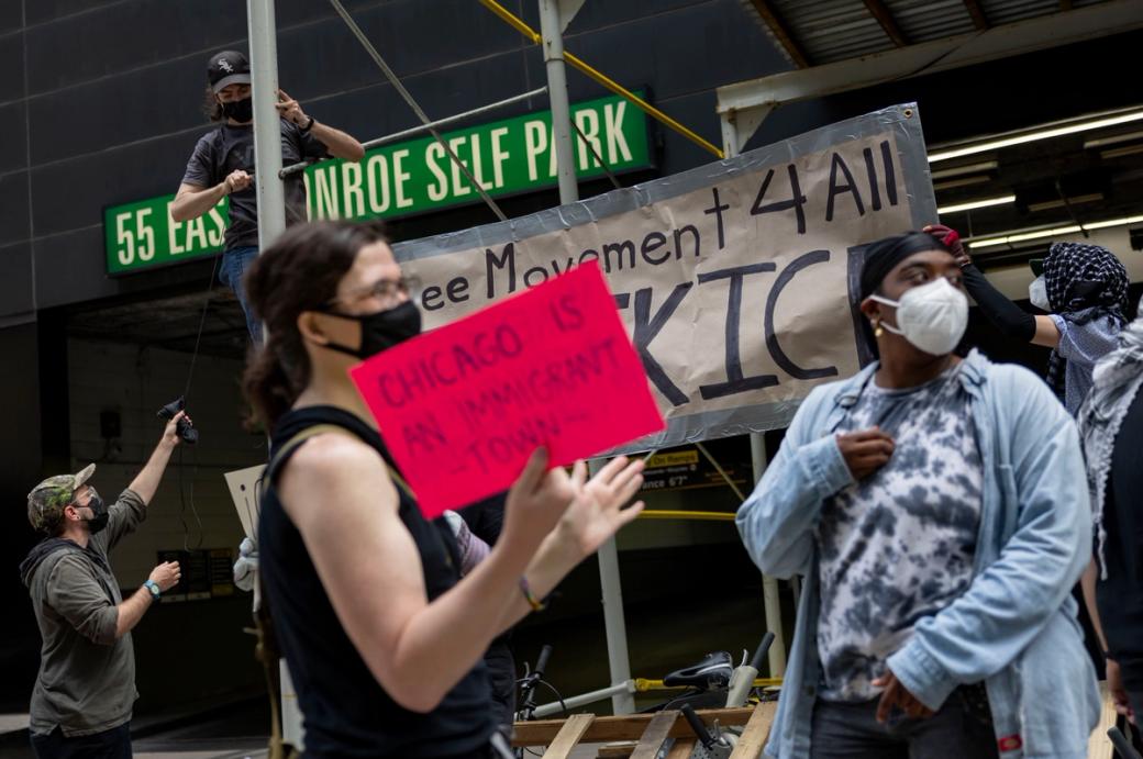 A group of people in masks in front of a sign that says 55 East Monroe Self Park. One person is holding a sign that says "Chicago Is An Immigrant Town"