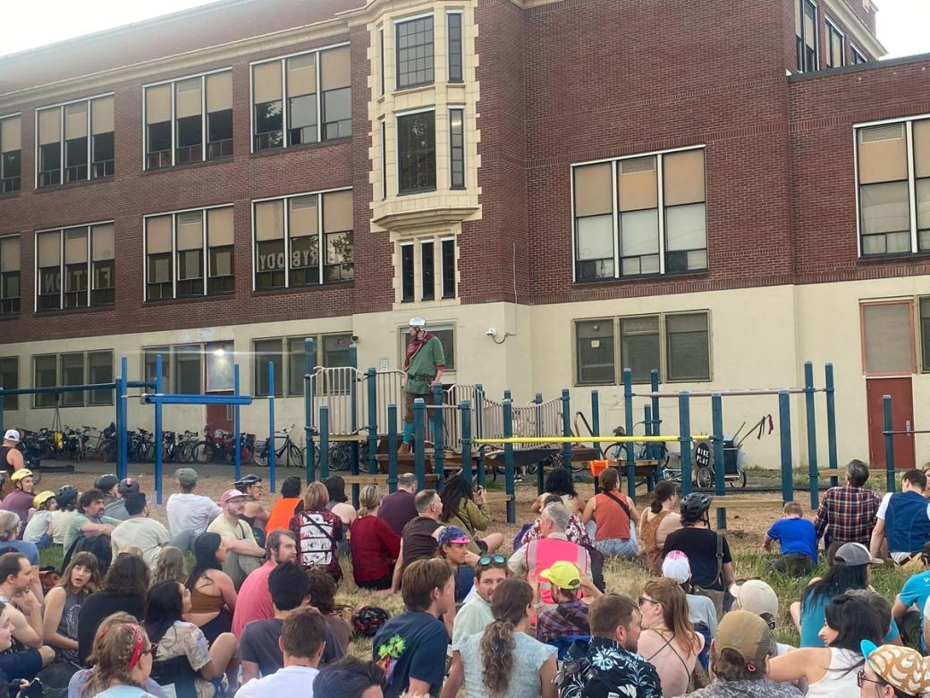 a crowd of people sitting in front of playground, large brick building in background