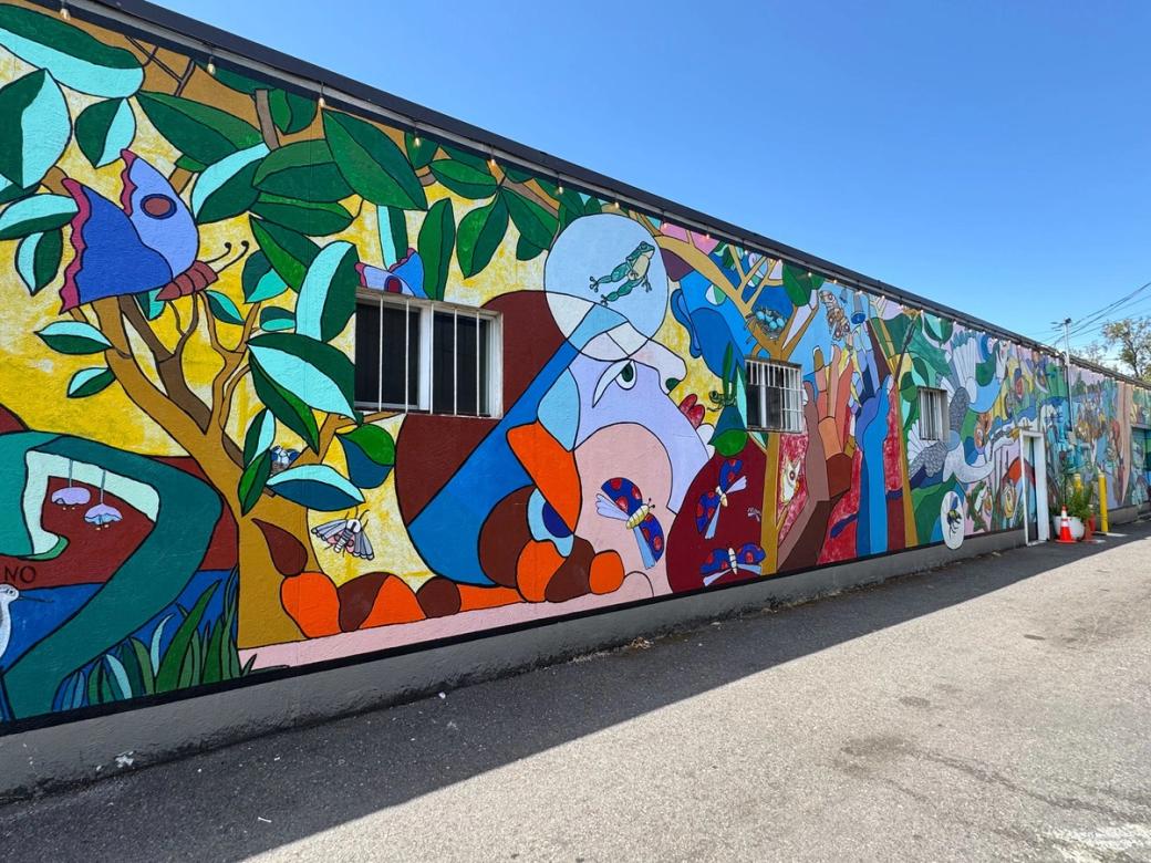 Brightly colored geometric shapes form a tree with a butterfly in the foreground on the long low wall with a mural.