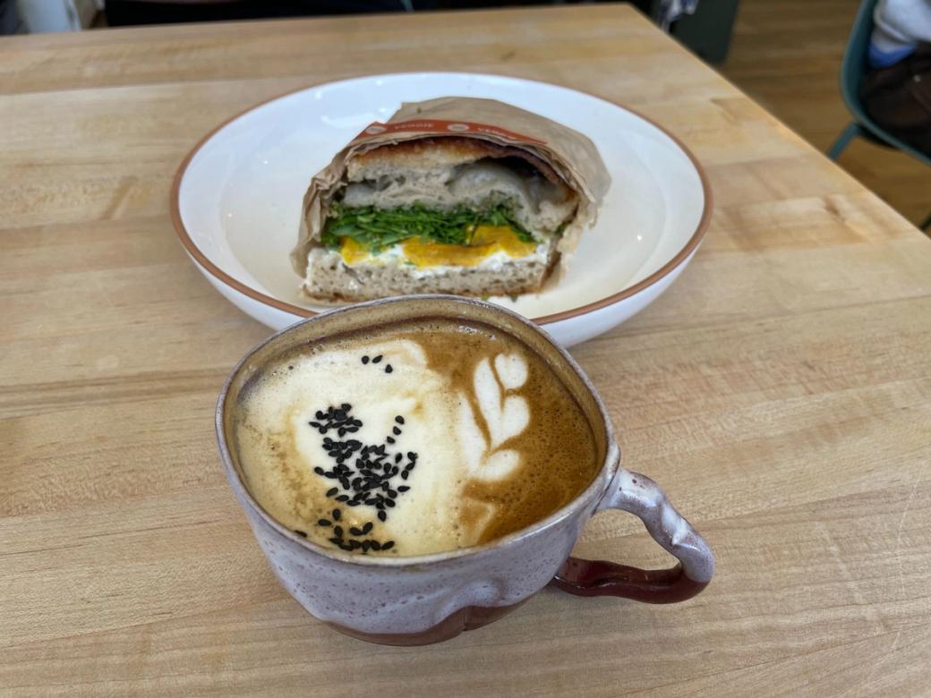 A ceramic coffee mug with design in the foam and sesame seeds on top sits on a cafe table with half a sandwich behind it.