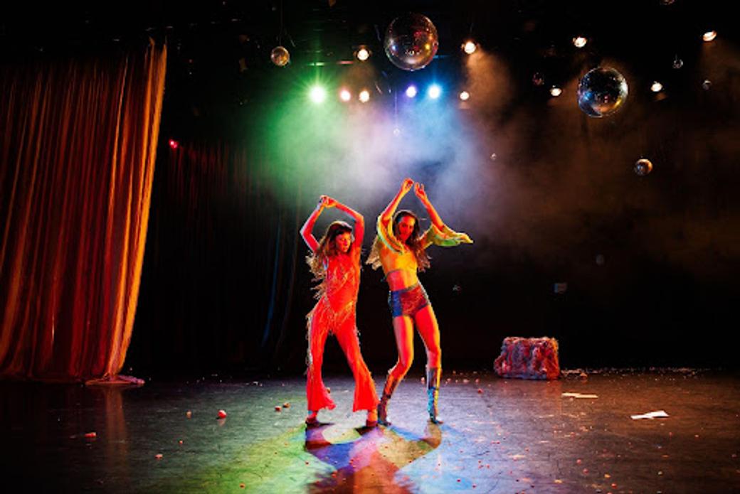 Two colorfully-dressed women in 70s-style clothing dancing on stage under multiple disco balls.