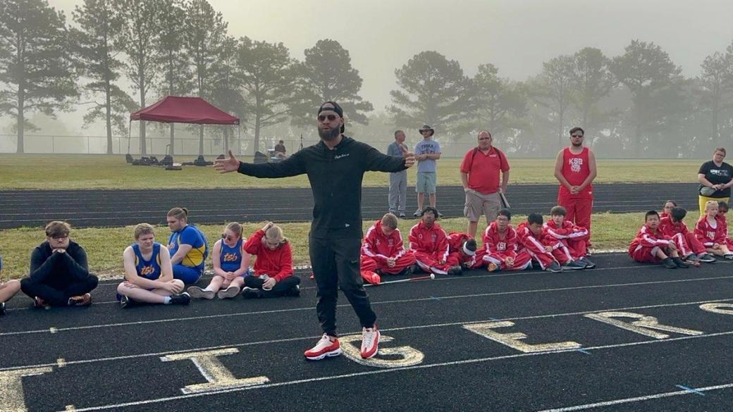 A Black albino man in a black track suit and hat speaks to kids on a track outside. The kids are seated on the track and wear blue and red track suits.