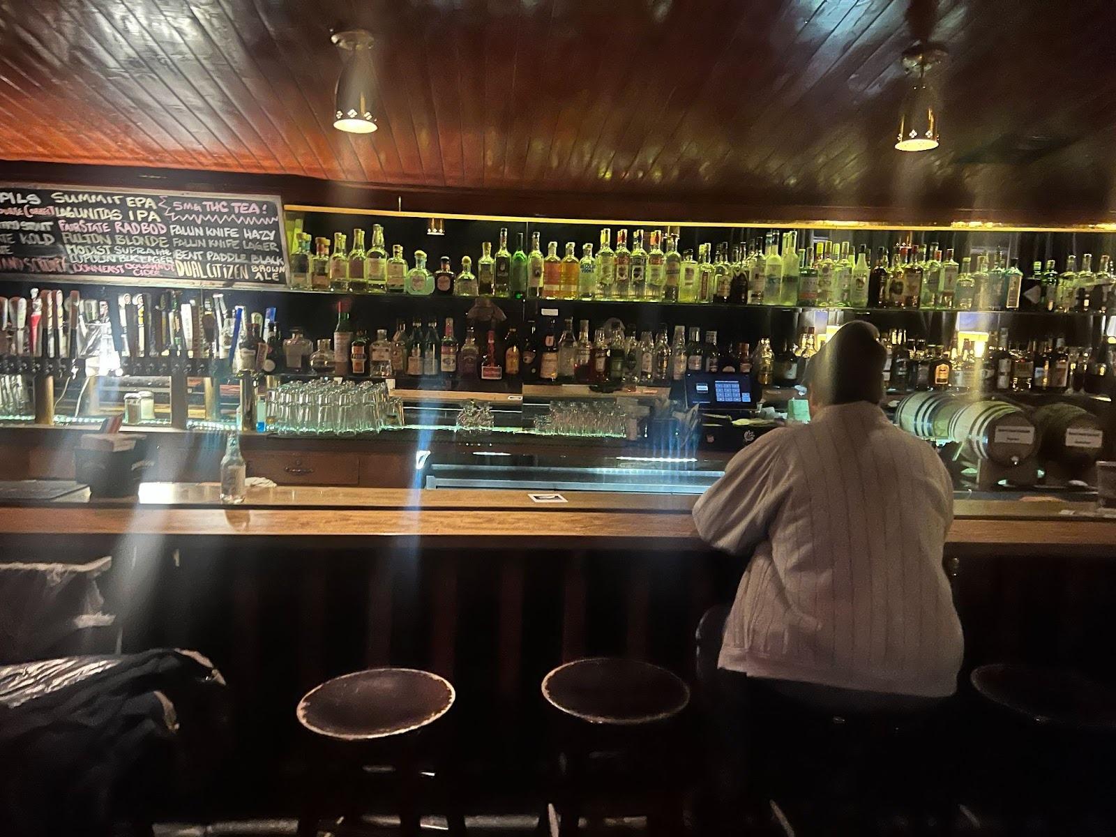 One person sitting at a bar. The bar is heavily stocked