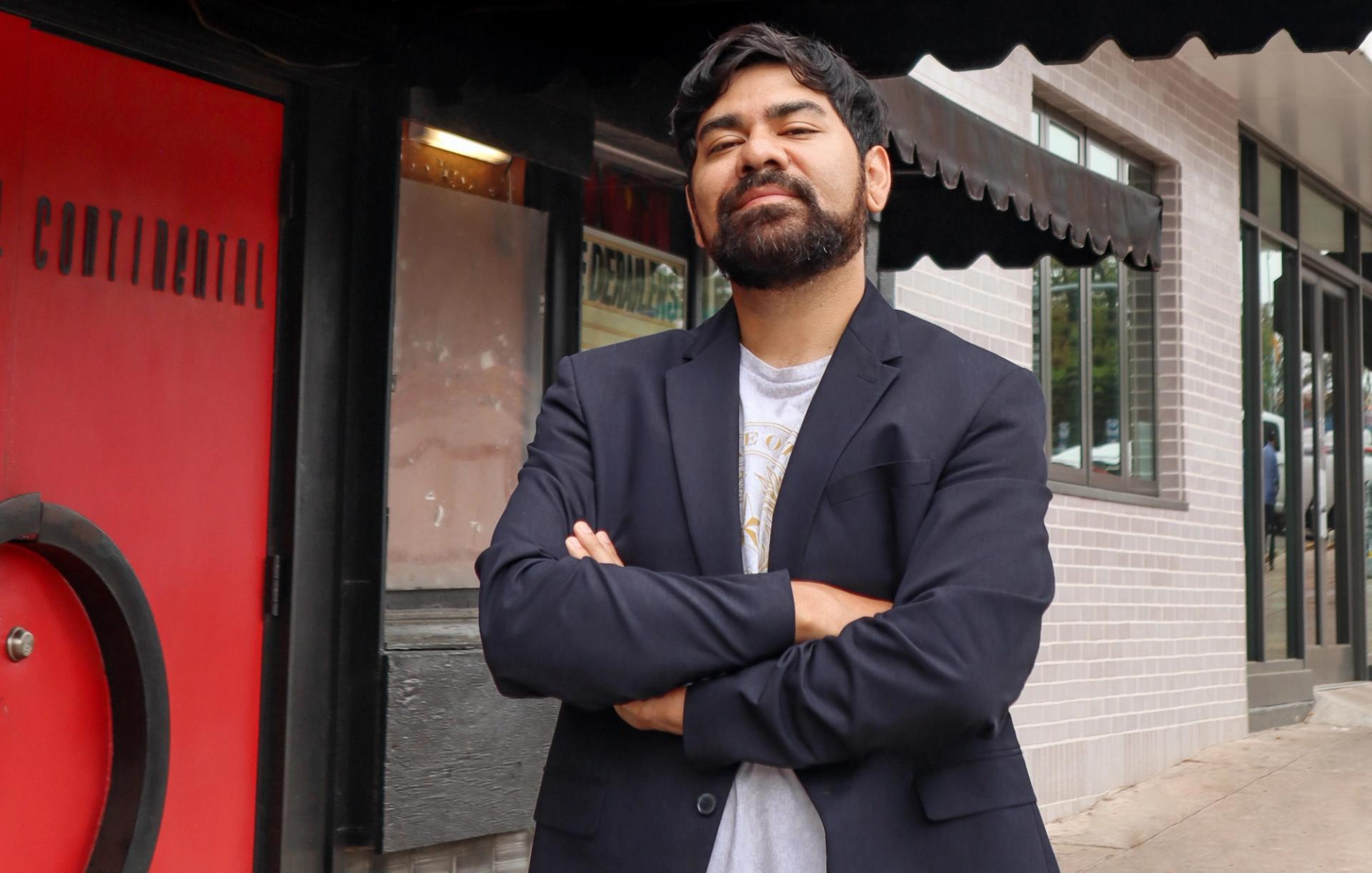 A man wearing a white shirt and a black blazer with his arms crossed. He is standing outside next to a red door.
