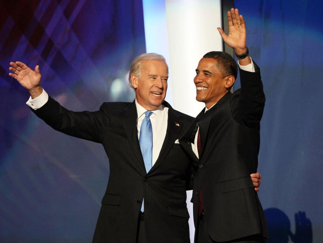 Joe Biden and Barack Obama stand on stage in suits waving at the crowd