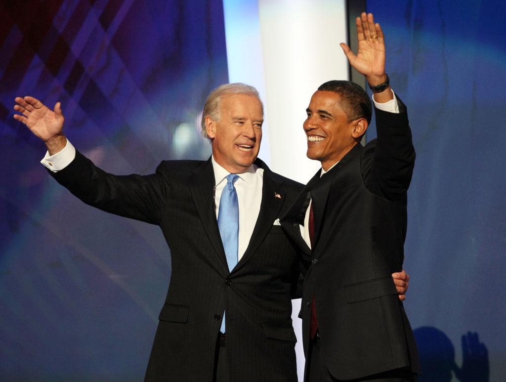 Joe Biden and Barack Obama stand on stage in suits waving at the crowd
