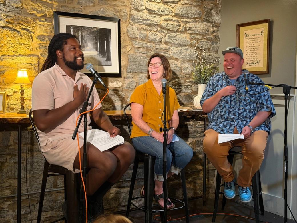 Three people sit on stools smiling and laughing, and holding microphones with stone wall behind them