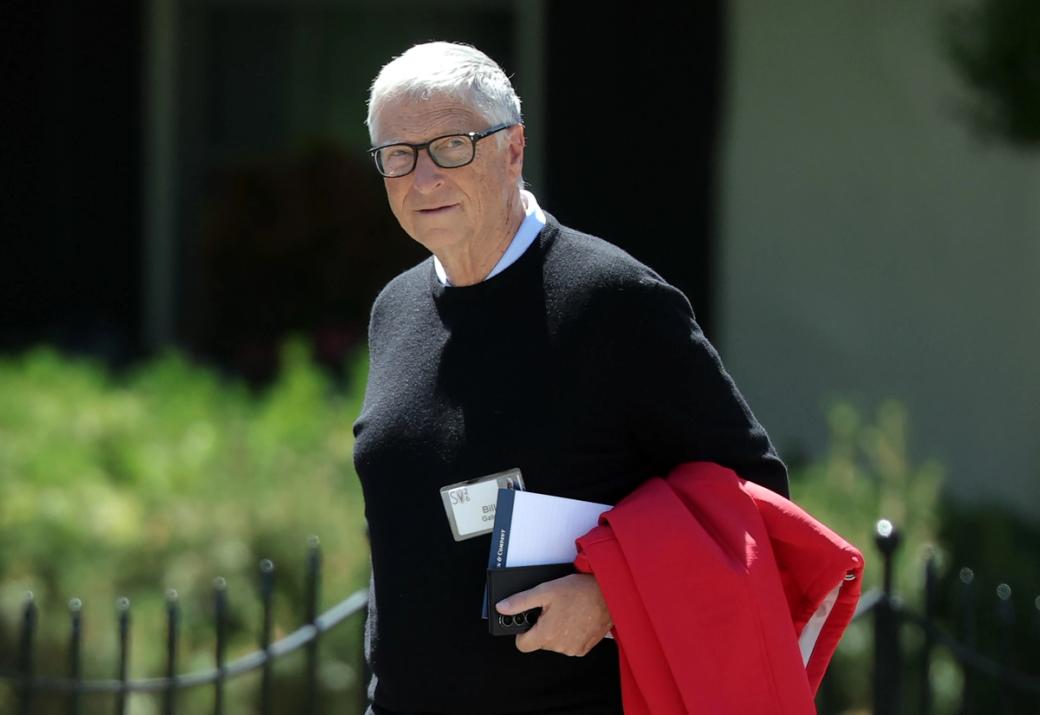 Bill Gates walks past the camera wearing a black sweater with a name tag pinned to it and carrying a paper pad and red jacket.