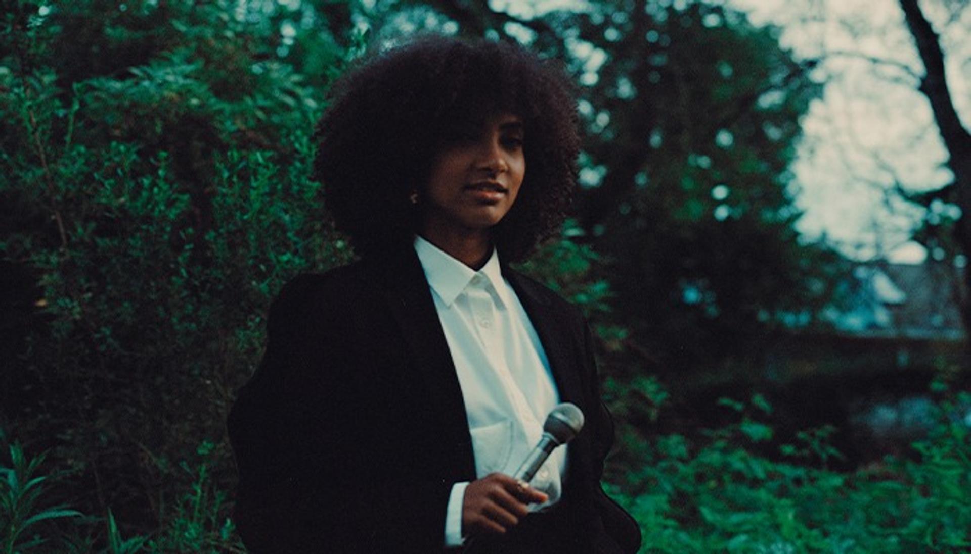 A woman in a lush field with trees. She's wearing a suit and holding a microphone