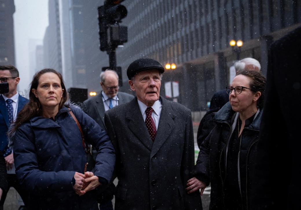 Man in suit stands outside on urban street, flanked by women on each side, and there is light snow falling