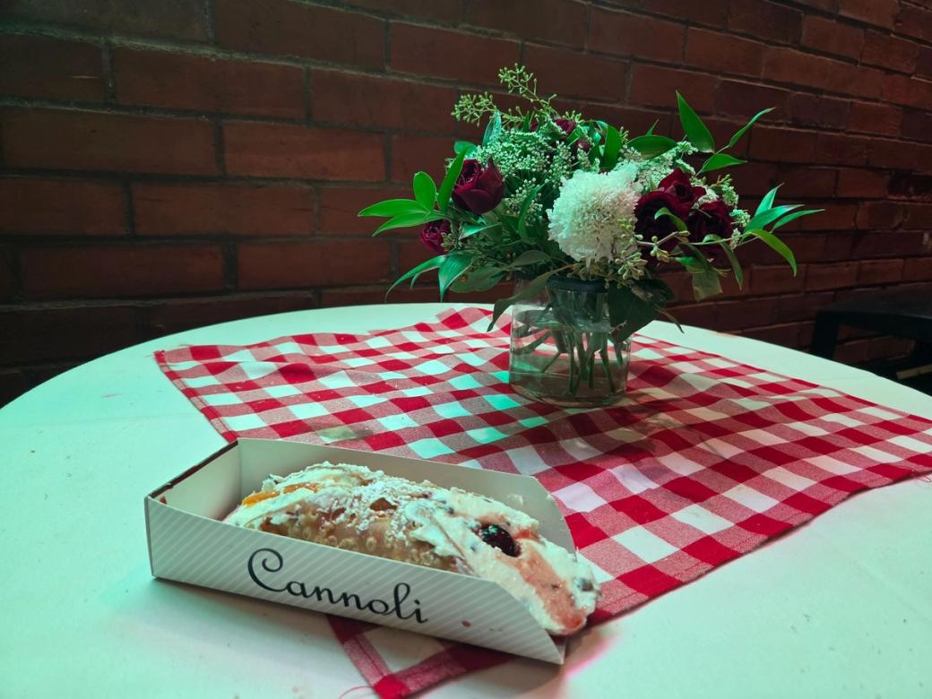 A cannoli in a holding box that says "cannoli" on a plaid table mat with a small bouquet of flowers in a vase