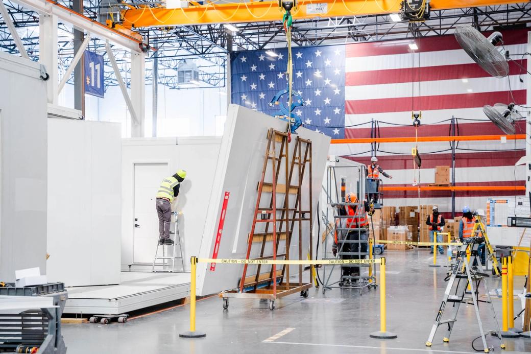 The inside of a warehouse with people in hard hats building things and a U.S. flag on the wall.