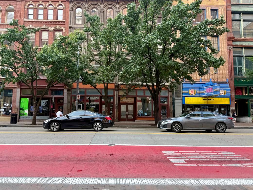 A wide view of a Downtown Pittsburgh street with a red-painted bus lane in the foreground, parked cars along the curb, leafy trees, historic brick buildings, and the Harris Theater marquee.