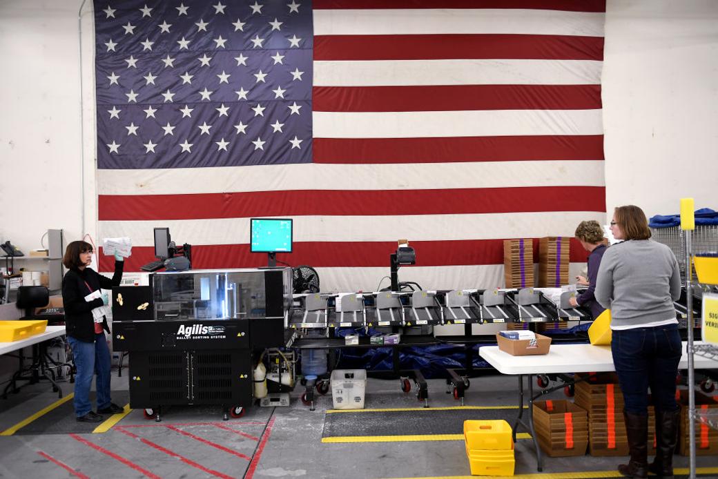 A ballot counting machine is manned by multiple election workers, with a large American flag hanging on the back wall.