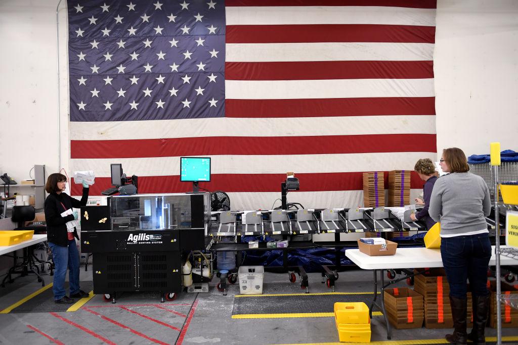 A ballot counting machine is manned by multiple election workers, with a large American flag hanging on the back wall.