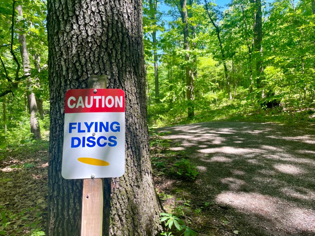 A sign that reads "Caution Flying Discs" on a tree along a wooded path