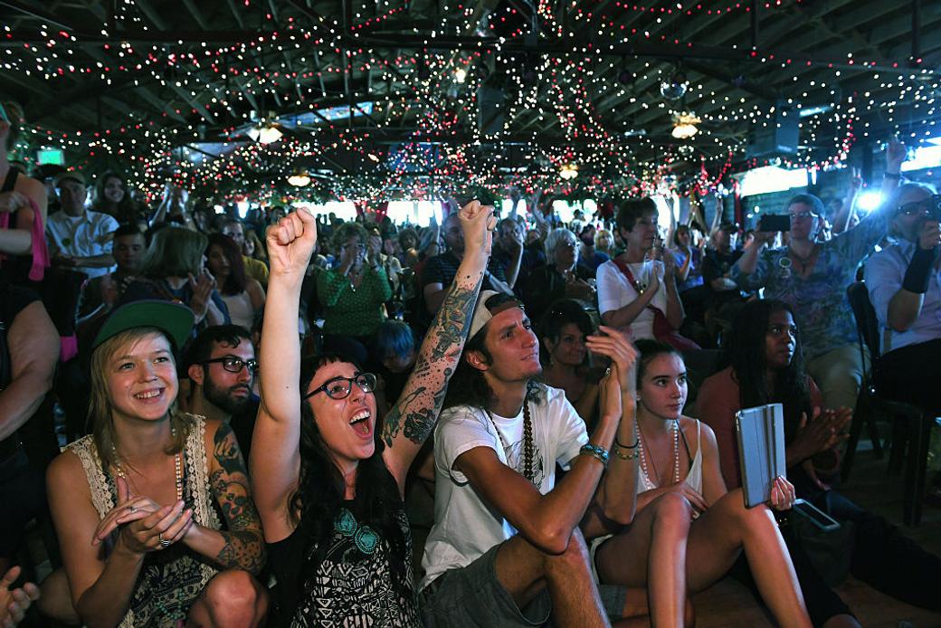 A person with glasses puts their fists up in the middle of a cheering crowd at the Mercury Cafe in 2016.