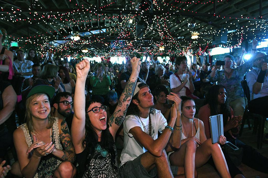 A person with glasses puts their fists up in the middle of a cheering crowd at the Mercury Cafe in 2016.