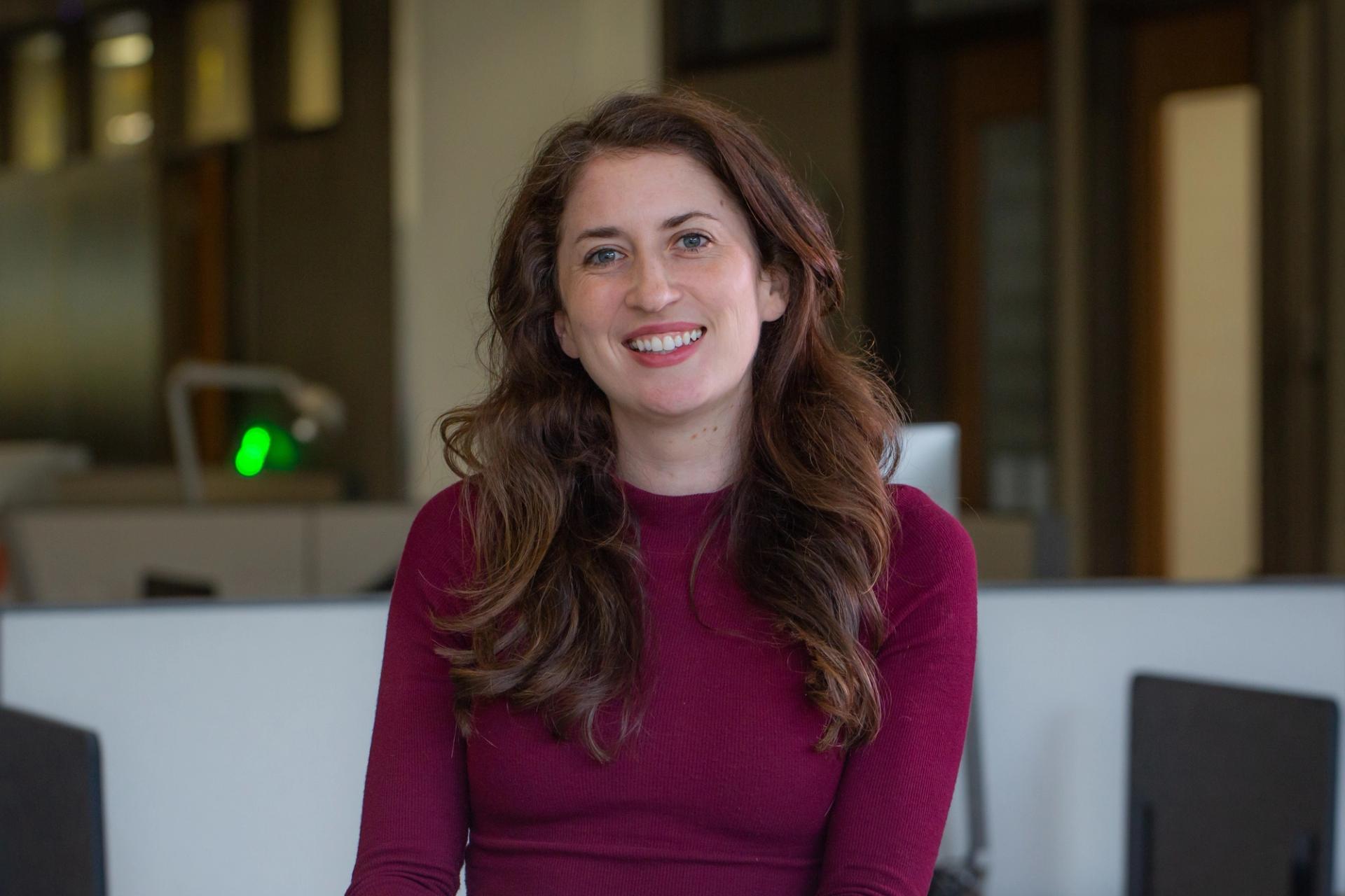 A woman with brownish, reddish hair wears a maroon long-sleeve shirt and smiles.