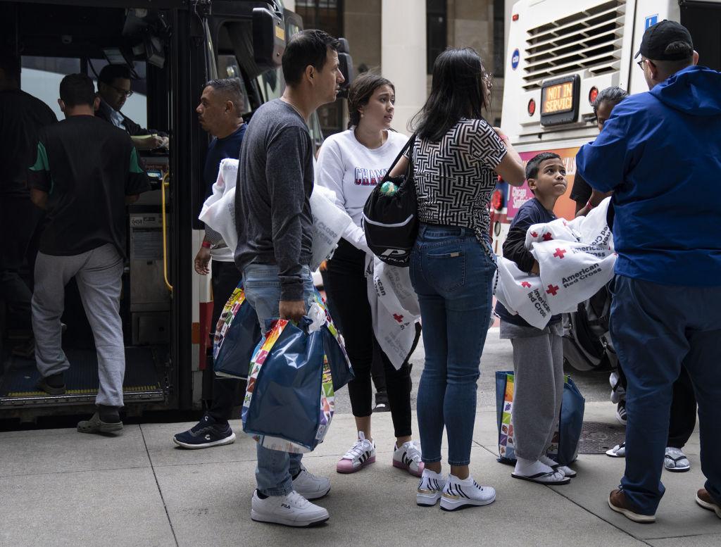 A group of migrants stands outside buses with bags and blankets