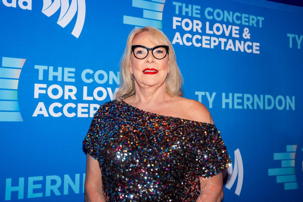 A white transgender woman with blonde hair and black glasses wears a sparkly dress in front of a blue background that says "The Concert for Love & Acceptance" and "Ty Herndon."