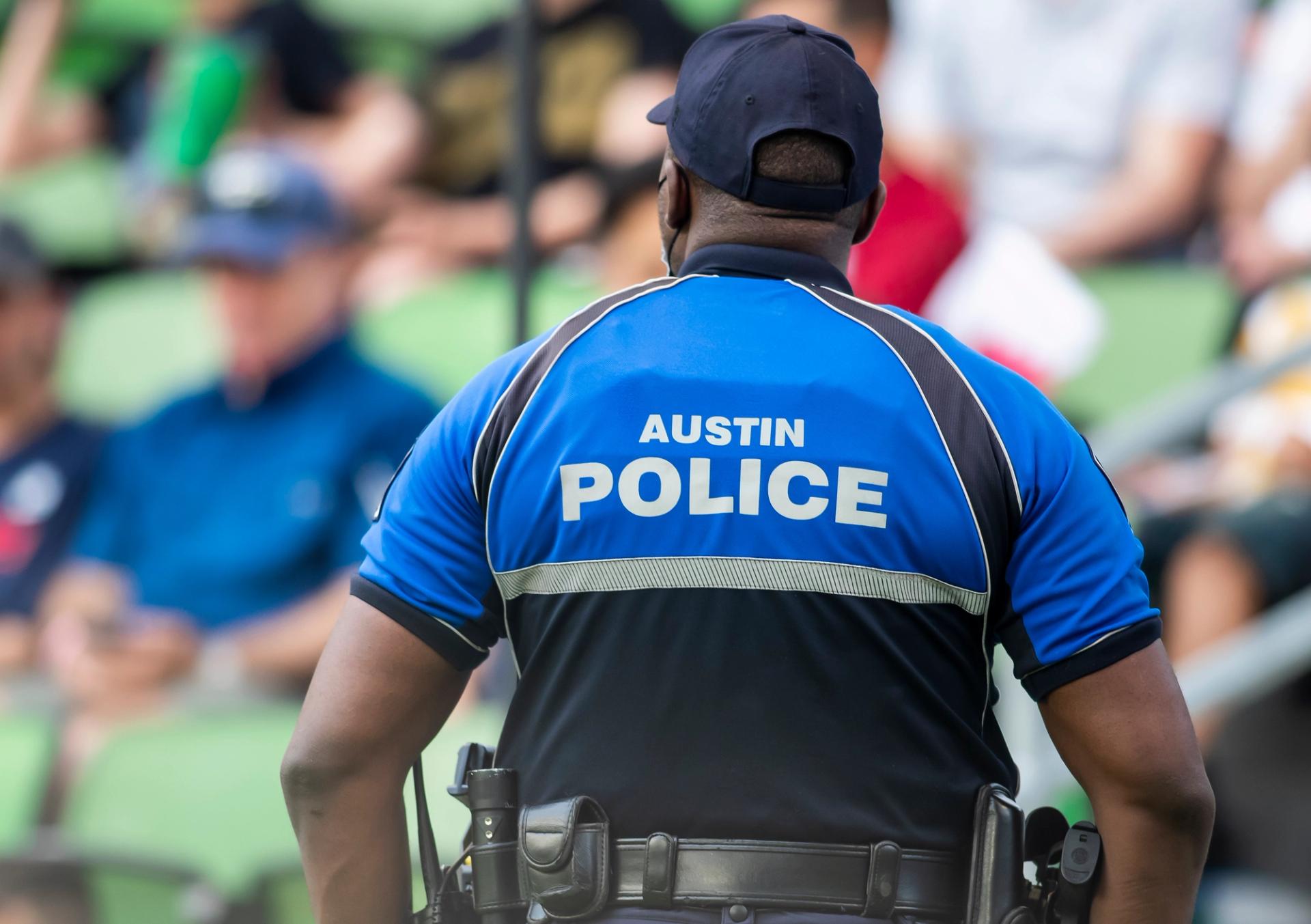 An Austin police officer's back. The officer is turned away from the camera and is wearing a black ball cap. A blue police uniform reads "Austin Police."