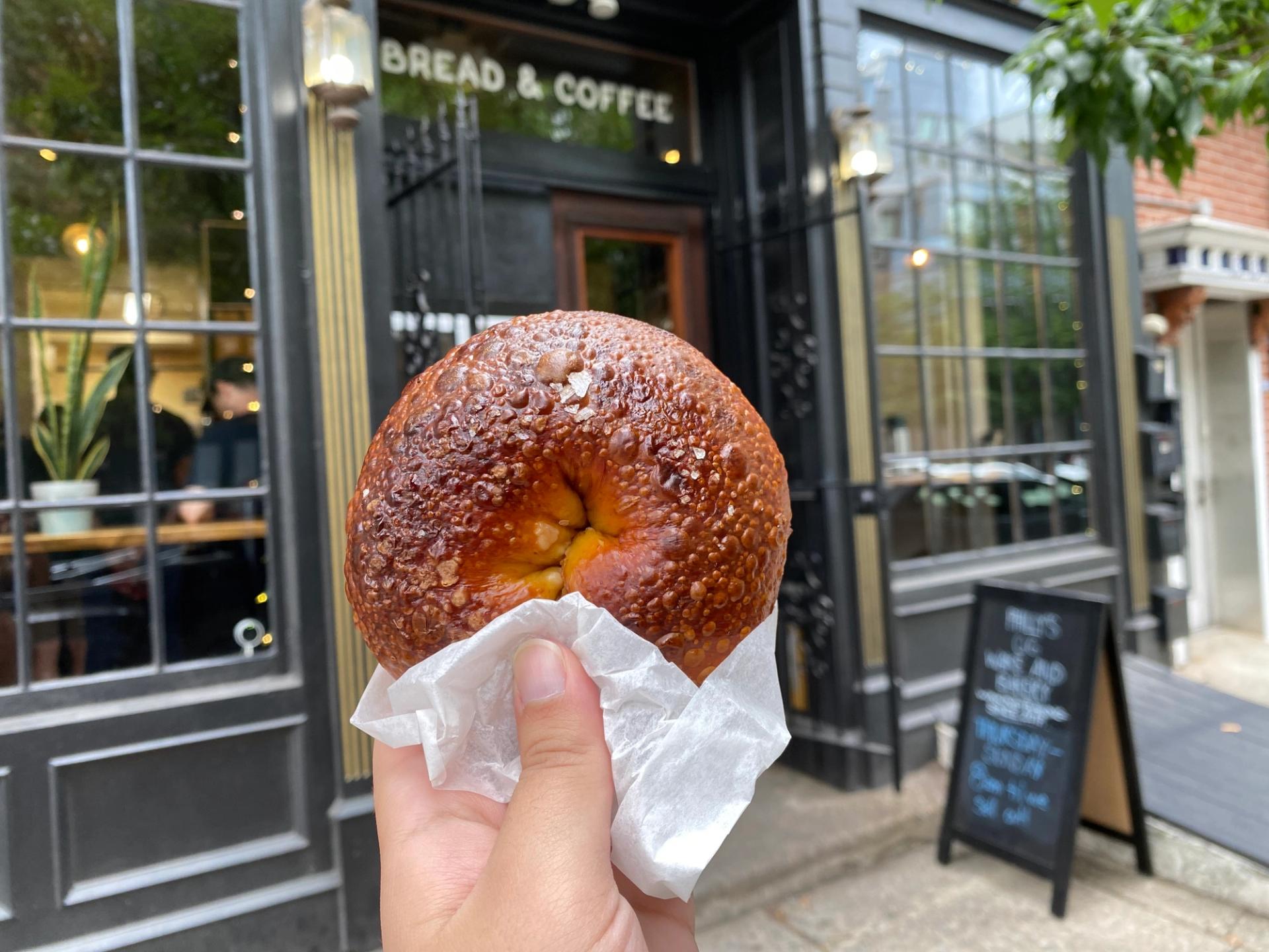 A pretzel bagel with The Kettle Black's storefront in the background.