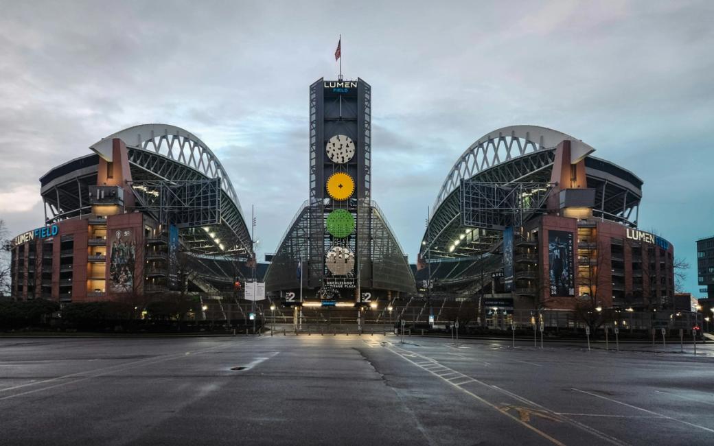 An exterior view of Lumen Field against a cloudy sky. The stadium has a distinctive curved roof and industrial design.