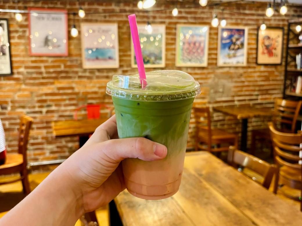 A hand holds an iced strawberry matcha latte layered in green and pink with a bright pink straw, inside a cozy café with exposed brick walls, string lights, and framed New Yorker magazine covers.