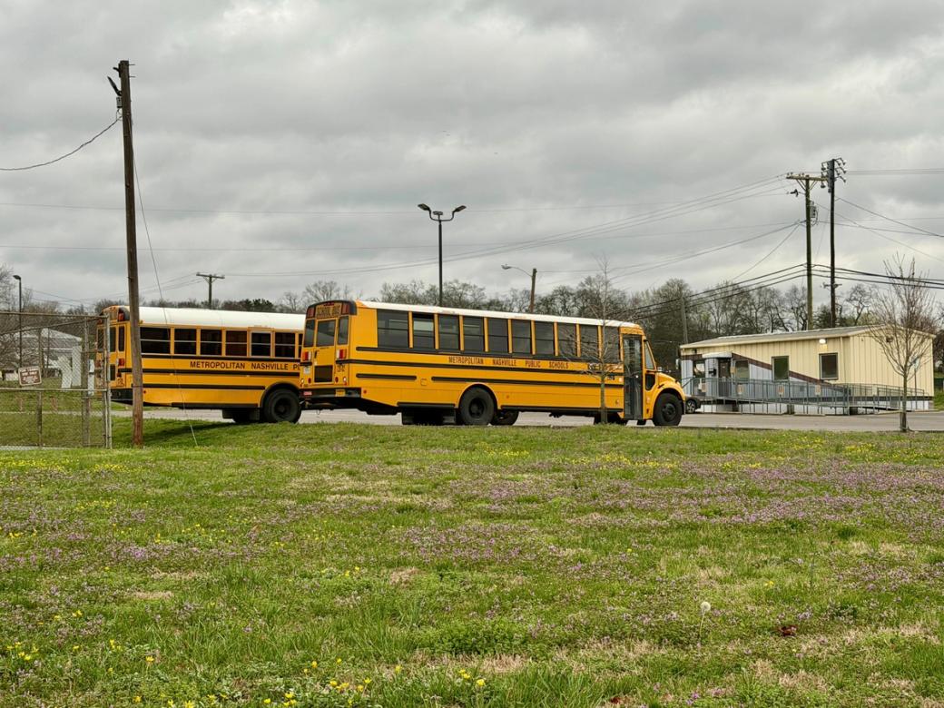 Two school buses in a parking lot beside a yellow and gray school trailer. The lot is surrounded by a grassy field with wildflowers.