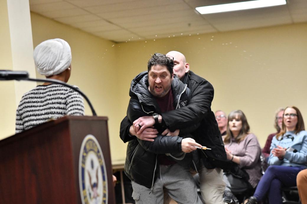Man restrained by another man attempting to lunge at woman standing next to podium, audience appears aghast in background
