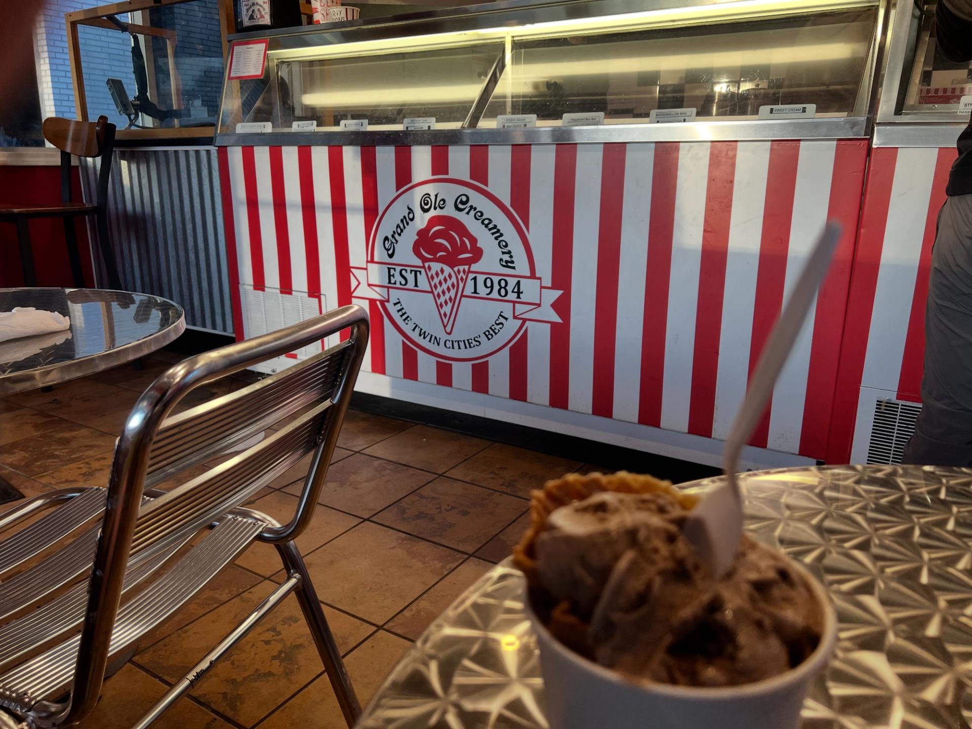 A photo of a cup of ice cream, in the back there is the serving area that reads "Grand Ole Creamery"
