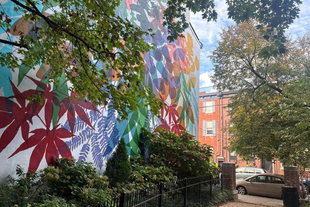 The side of a rowhome is covered in a mural depicting abstract leaves in red, yellow, green, purple, orange, and blue. The mural faces a small park bordered by a wrought iron gate. The entrance to the park has two small brick pillars. The branches of trees outline the top, right, and left sides of the photograph. 