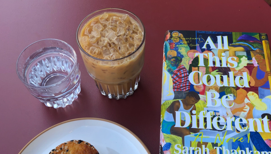 An ice latte in a clear glass, next to a glass of water, a pastry on a plate and a book with the title “All This Could Be Different.”