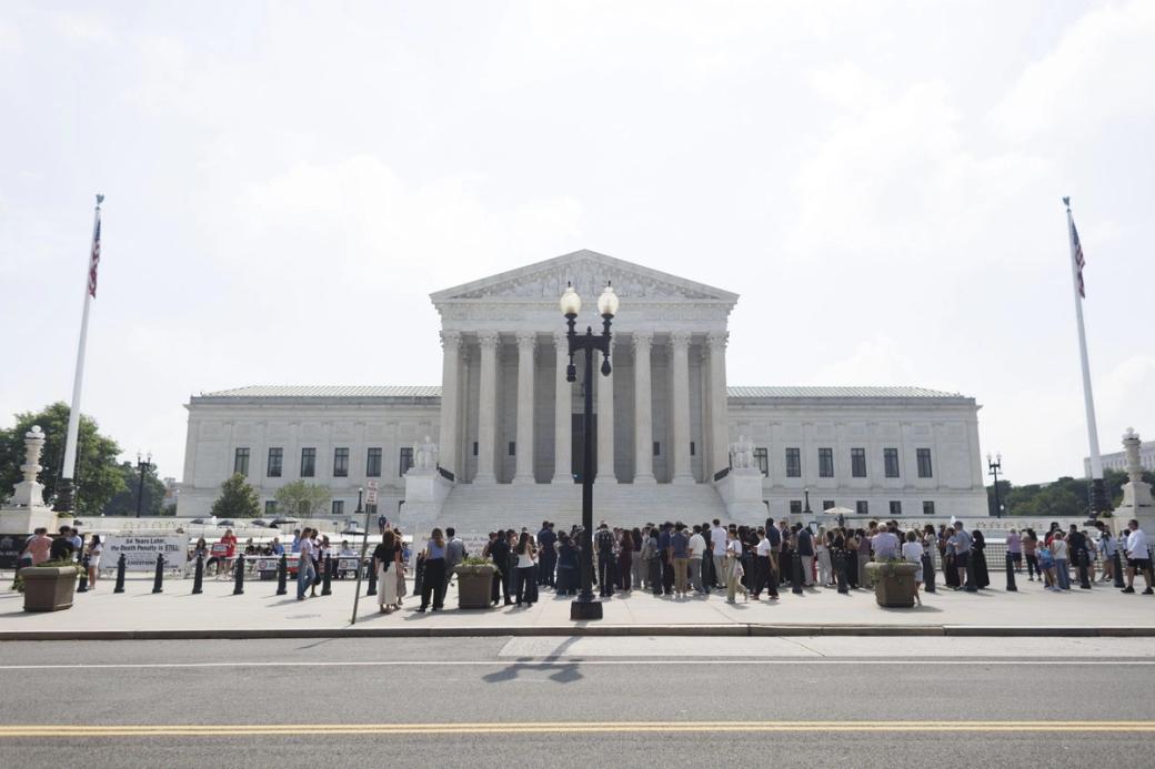 A street view of a large white pillared building. There are a group of people gathered outside of it as well