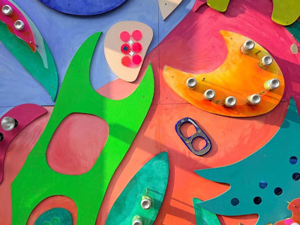 Flat sheets of wood painted bright, primary colors are arranged against a two-tone backdrop, with soda bottles and other recyclables mounted on the wood.