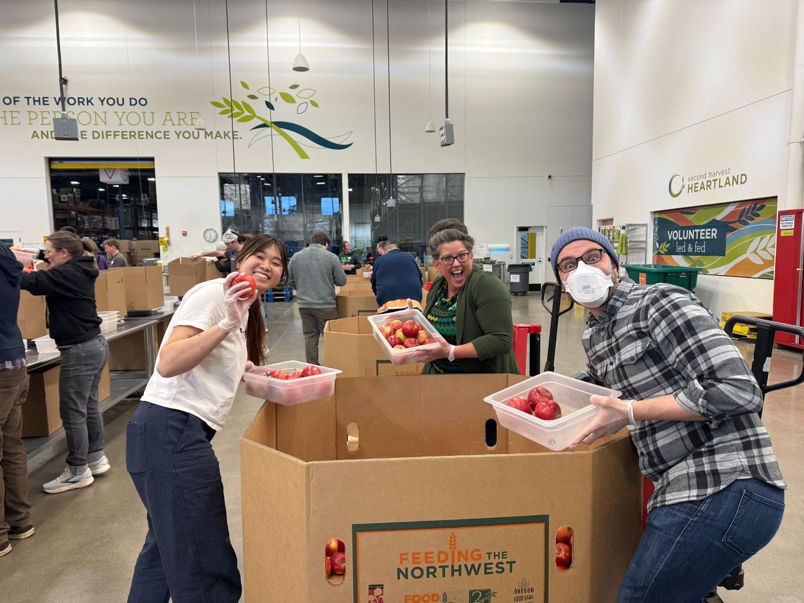 A group of people posing for a photo. They are gathered around a cardboard box filled with apples. They are showing off their plastic tub with apples in it