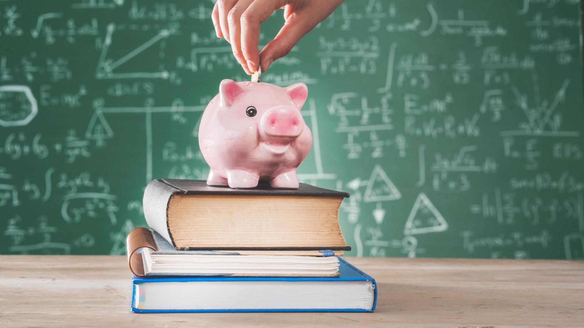 A piggy bank on top of two books. A chalkboard is in the background with math formulas on it. A person is pointing a coin into the piggy bank.