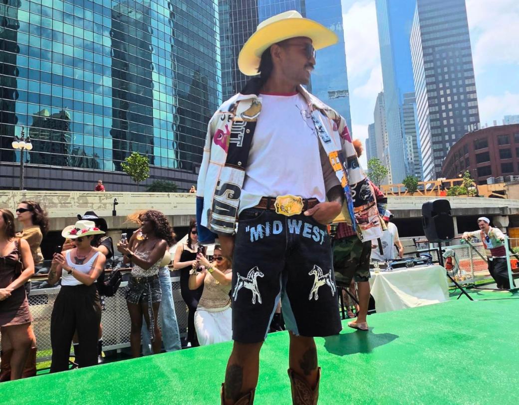 A man in a cowboy hat, white t shirt, pants that say "mid west" and wearing a belt with a huge belt buckle. He is standing on a green platform