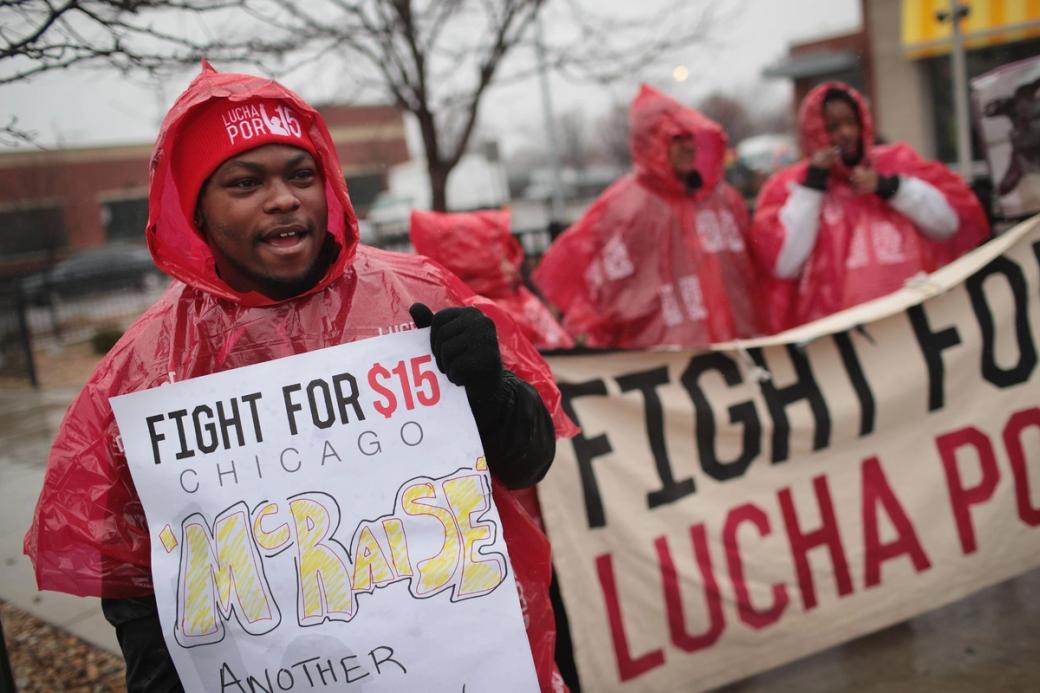 A group of people in red rain jackets protesting. They have signs that are demanding a raise to $15
