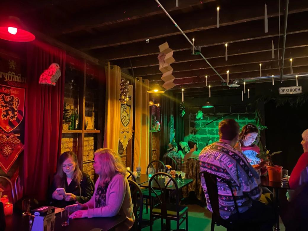 The interior of the Harry Potter pop-up bar featuring floating candles; red, yellow, and green Hogwarts house banners and curtains; and a brick wall in the background. Guests sit at tables around the 